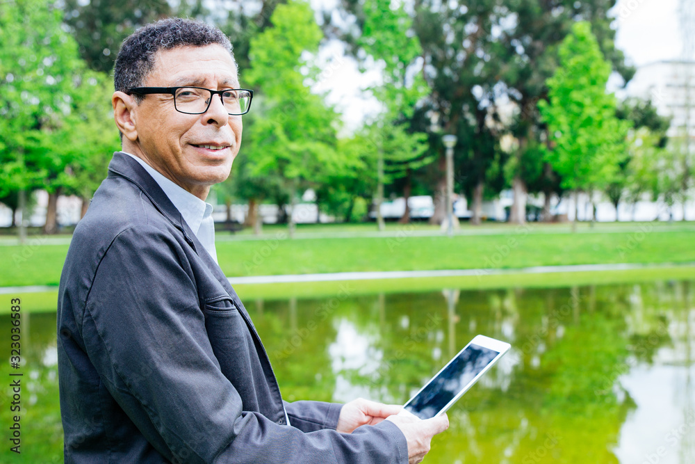 Smiling man using tablet and standing in city park. Guy wearing casual clothes and holding gadget with pond and green trees in background. Communication and nature concept. Side view. Smiling man using tablet and standing in city park. Guy wearing casual clothes and holding gadget with pond and green trees in background. Communication and nature concept. Side view.