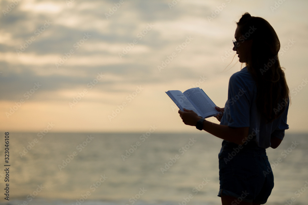 a silhouette of a beautiful girl in denim shorts and a blue shirt stands near the sea and reads a book at sunset
