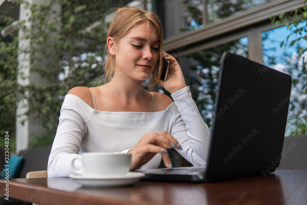 Woman working on laptop at office while talking on phone, backlit warm light. Portrait of young smiling business woman calling her best friend, having break, telling something funny, sitting in cafe. Woman working on laptop at office while talking on phone, backlit warm light. Portrait of young smiling business woman calling her best friend, having break, telling something funny, sitting in cafe.