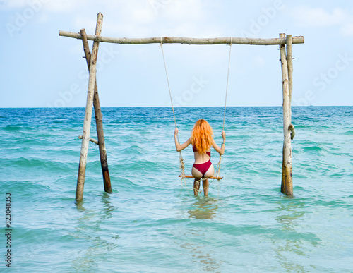 Beach summer vacation swing on th sea with young beautiful girl sitting on beach swing blue sea.Red haired woman swinging on the beach on Phu Quoc island, Vietnam. Happy on tropical ocean swing.