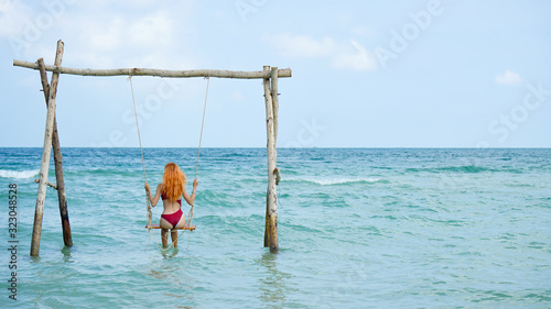 Beach summer vacation swing on th sea with young beautiful girl sitting on beach swing blue sea.Red haired woman swinging on the beach on Phu Quoc island, Vietnam. Happy on tropical ocean swing.
