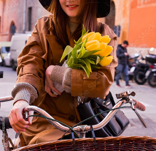 Beautiful girl in a raincoat riding a bicycle with with a vintage basket holds a bouquet of yellow flowers. Walks on the city of Europa. Romantic and happy mood girls.