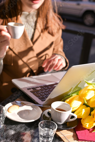 A young girl drinks coffee in a cafe on the street and works on the Internet on a laptop. Sunlight in the frame. Traveling and walking with a noutbuk. Copy space. photo