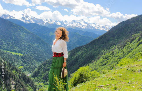 Elegant woman in green skirt at the mountains. Happy girl with wavy hair with a hat. The background of the majestic mountains of the Caucasus and wooden fence. Svaneti, Georgia.