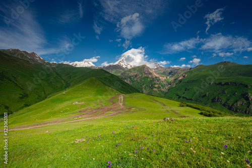 Mount Kazbek in Georgia (Caucasus) and green meadows at the foot of the mountain. The top of the mountain is obscured by clouds and fog on a sunny and clear day.