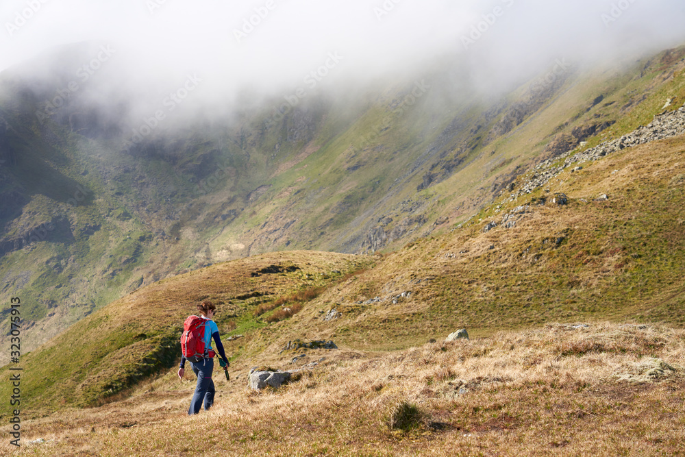 A female hiker and their dog walking up hill towards the mountain summit of Beda Fell with Heck Cove to the left on a sunny day in the Lake District UK.
