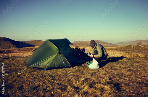 A female hiker preparing to make a hot drink beside their tent that s pitched on a mountain summit at sunrise at Rampsgill Head in the Lake District UK..