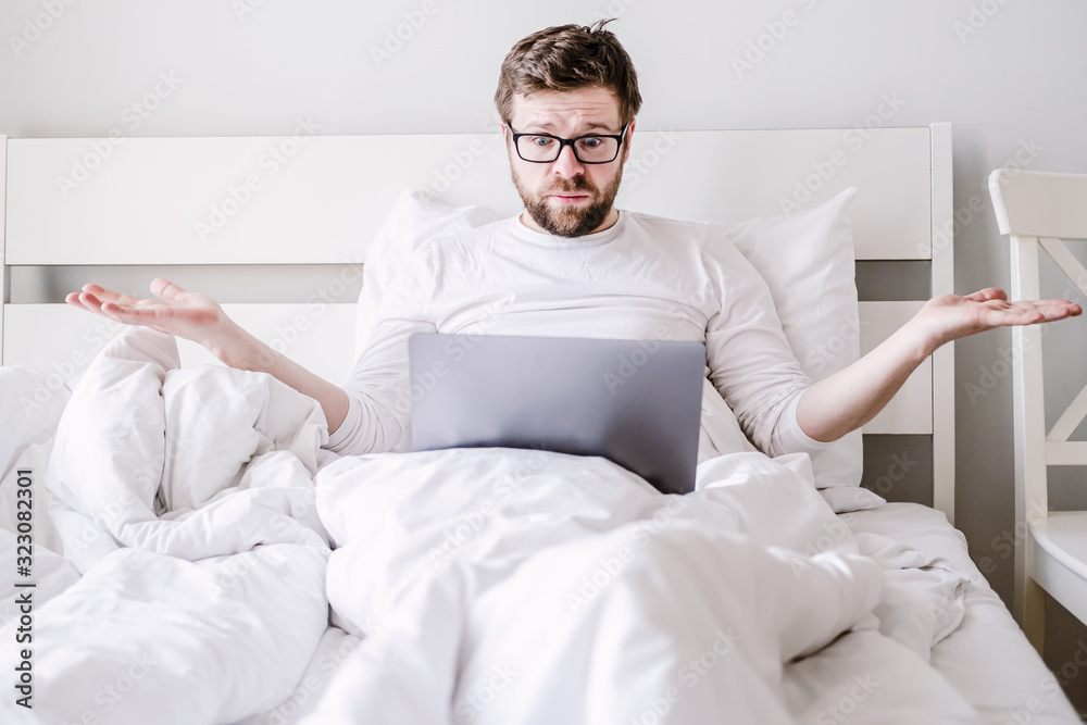 Upset man in pajamas in bed with a laptop, he stares at the screen in perplexity and makes hands to the sides.