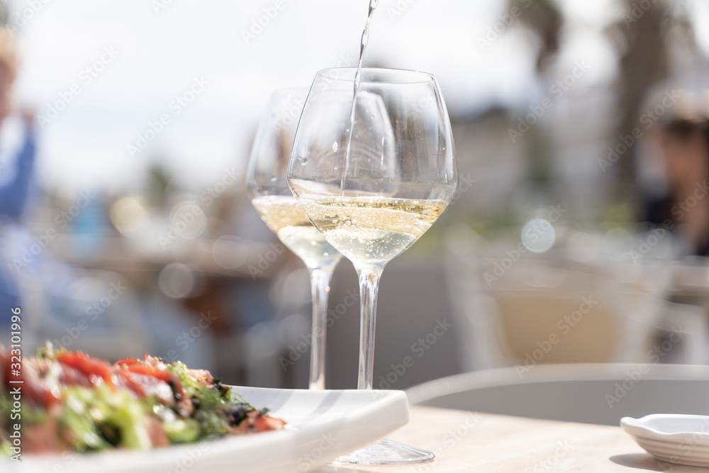 Detail of two glasses of white wine while filling one of them and a rectangular plate with smoked salmon and salad with seeds on top at a restaurant table Detail of two glasses of white wine while filling one of them and a rectangular plate with smoked salmon and salad with seeds on top at a restaurant table