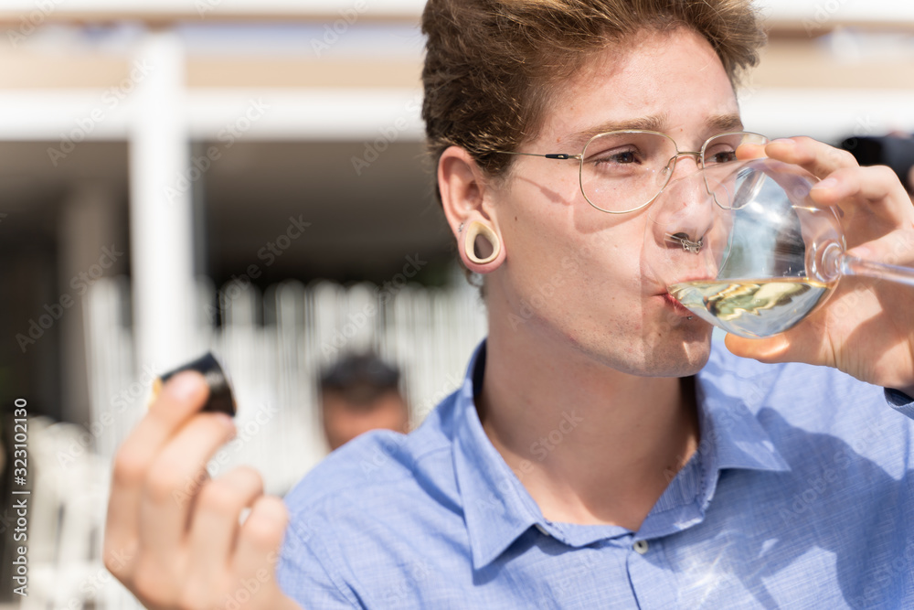 young man with piercings and lasses drinking white wine while is holding a piece of sushi in the other hand young man with piercings and lasses drinking white wine while is holding a piece of sushi in the other hand