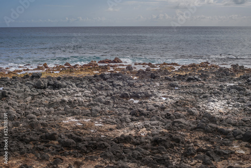 Kauai, Hawaii, USA. - January 11, 2012: Black lava rocks at Kawailoa beach in front of azure and gray ocean under light blue sky with far away clouds. photo