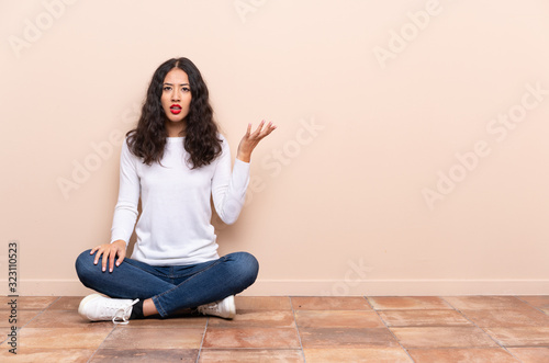 Young woman sitting on the floor making doubts gesture