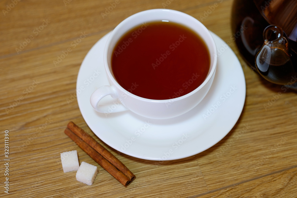 Classic white cup of black tea with saucer, teapot, sugar and cinnamon on the wooden table background. Breakfast drink at home or cafe. Classic white cup of black tea with saucer, teapot, sugar and cinnamon on the wooden table background. Breakfast drink at home or cafe.