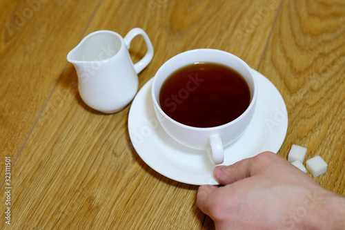 Male hand holding white cup with tea, sugar and milk teapot on the wooden table top view.