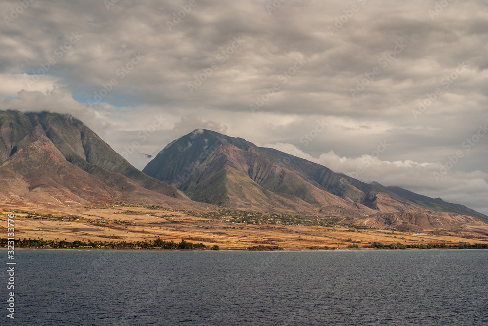Lahaina, Maui, Hawaii, USA. - January 12 2012: West coast dark ocean shoreline shows brown-yellow dry meadows, a town and 2 dark gray-brown mountains under massive gray cloudscape. Lahaina, Maui, Hawaii, USA. - January 12 2012: West coast dark ocean shoreline shows brown-yellow dry meadows, a town and 2 dark gray-brown mountains under massive gray cloudscape.