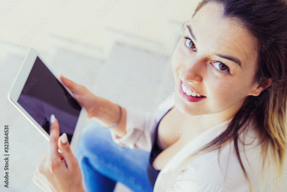 Cheerful tablet user browsing internet outside. High angle of young woman in white jacket sitting on outdoor stair, holding mobile pc and smiling at camera. Wi-Fi connection concept Cheerful tablet user browsing internet outside. High angle of young woman in white jacket sitting on outdoor stair, holding mobile pc and smiling at camera. Wi-Fi connection concept