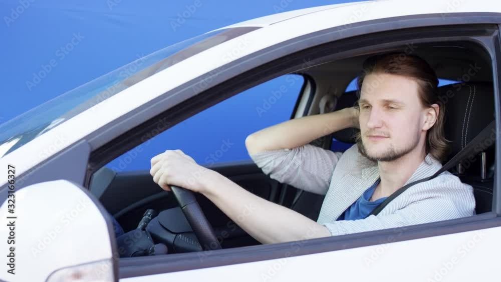 Complacent man is looking himself in the outside car mirror. Self-satisfied young guy is stroking his hair and singing a song in a car. Cheerful driver is sitting in white car against blue background.