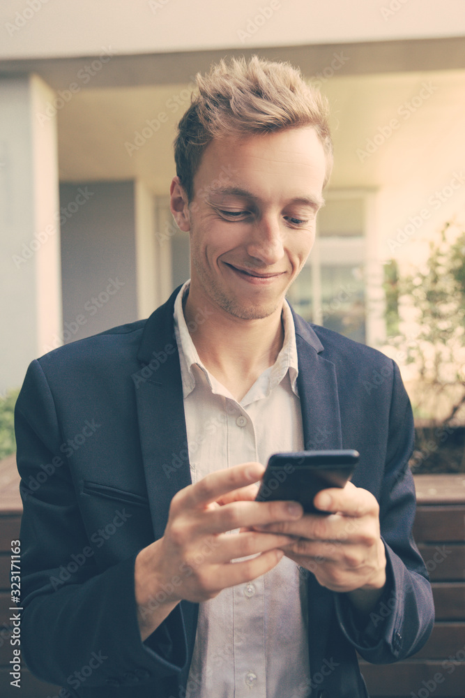 Happy pensive businessman texting message on phone outside. Handsome young man in office suit using cell, looking at screen and smiling. Communication concept