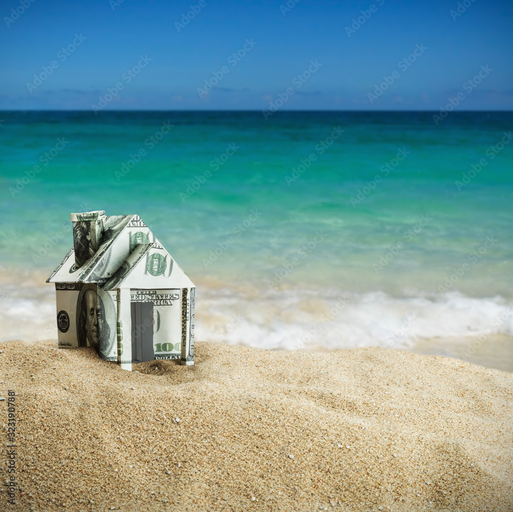 Naklejka premium house cut out of dollar bills on the sand. Concept on the topic of selling or buying a home.