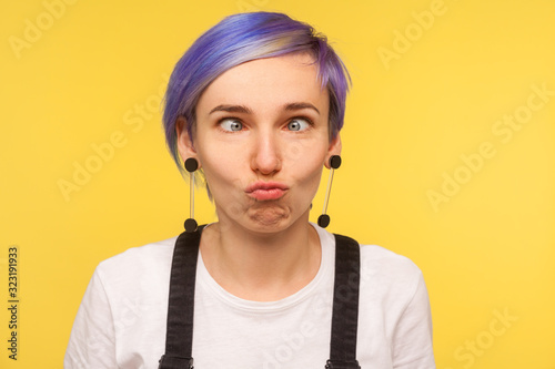 Portrait of comic hipster girl with violet short hair looking cross eyed, making silly awkward face expression, having fun and playing fool. isolated on yellow background, indoor studio shot, closeup photo