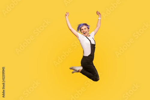 Portrait of ecstatic lively energetic hipster girl with violet hair in overalls jumping in air celebrating victory, personal success, rejoicing big sale. isolated on yellow background, studio shot
