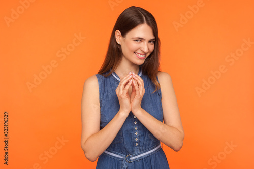 Stunning sly brunette woman in denim dress thinking over mysterious tricky plan, smirking with cunning playful expression, scheming evil plan, devious idea. studio shot isolated on orange background photo