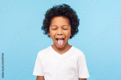 Portrait of funny naughty little boy with curly hair in T-shirt sticking out tongue and keeping eyes closed, disobedient child foolishing with cute derisive expression. studio shot blue background