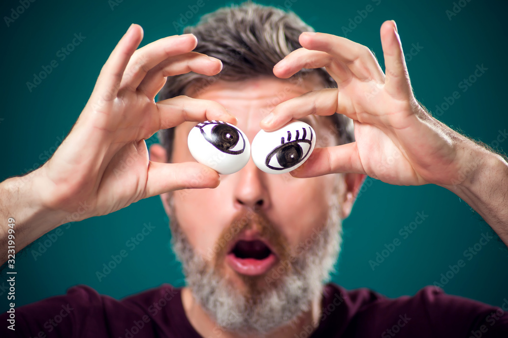 A portrait of funny bearded man holding eggs behind eyes. People, emotions and food concept A portrait of funny bearded man holding eggs behind eyes. People, emotions and food concept