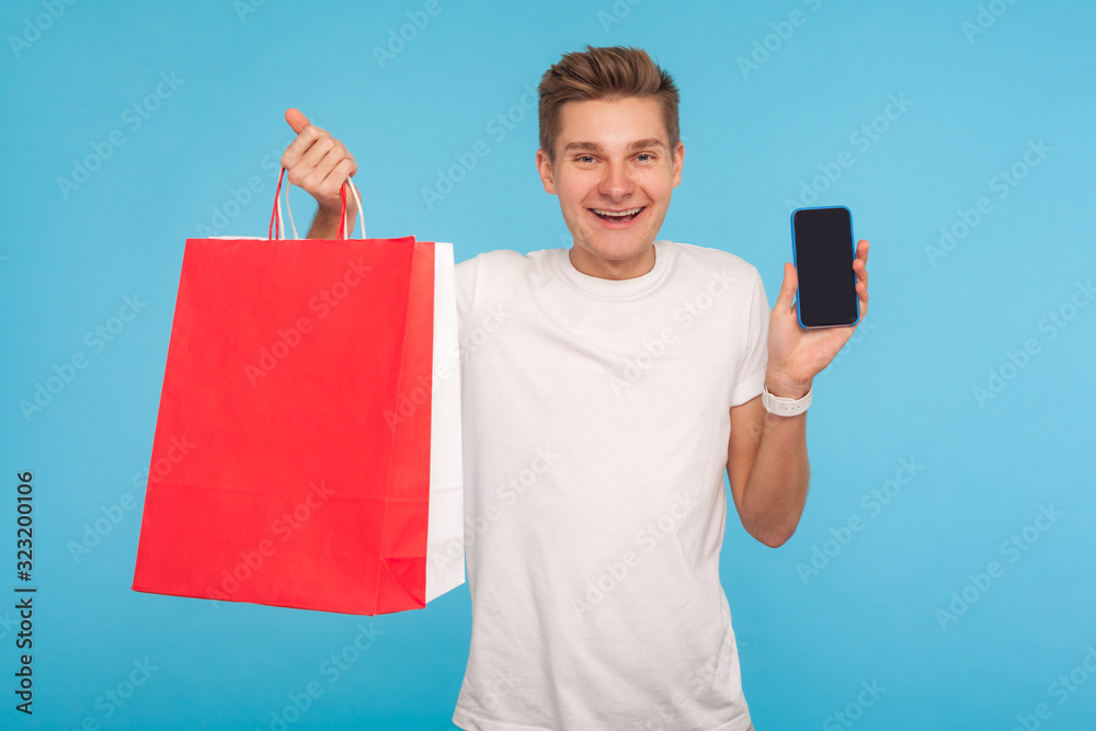 Mobile app for online shopping. Joyful happy man in casual white t-shirt holding cell phone and packages, smiling at camera, advertising of online store. indoor studio shot isolated on blue background Mobile app for online shopping. Joyful happy man in casual white t-shirt holding cell phone and packages, smiling at camera, advertising of online store. indoor studio shot isolated on blue background