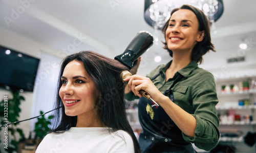 Girl’s day. Close-up photo of a smiling hairdresser drying and straightening hair of her gorgeous customer.