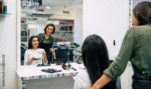 A small talk. Young hairstylist with a round comb and her beautiful female customer with a coffee cup are looking in the mirror and smiling.