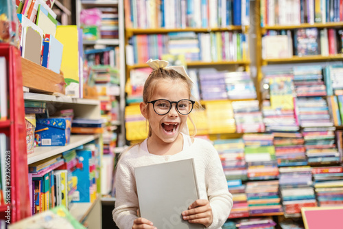 Excited caucasian cute little blond girl with eyeglasses and ponytail standing in bookstore and holding book she was looking for.