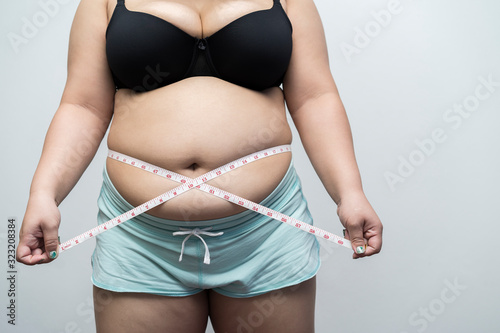 young asian woman wearing a bra and wearing light blue shorts check out her body fat while holding measuring tape around waist on gray background. concept obesity, exercise,overweight or body firming