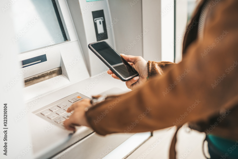 Close up shot of young adult woman. He typing pin code on keypad of ATM machine while using smart phone. Close up shot of young adult woman. He typing pin code on keypad of ATM machine while using smart phone.
