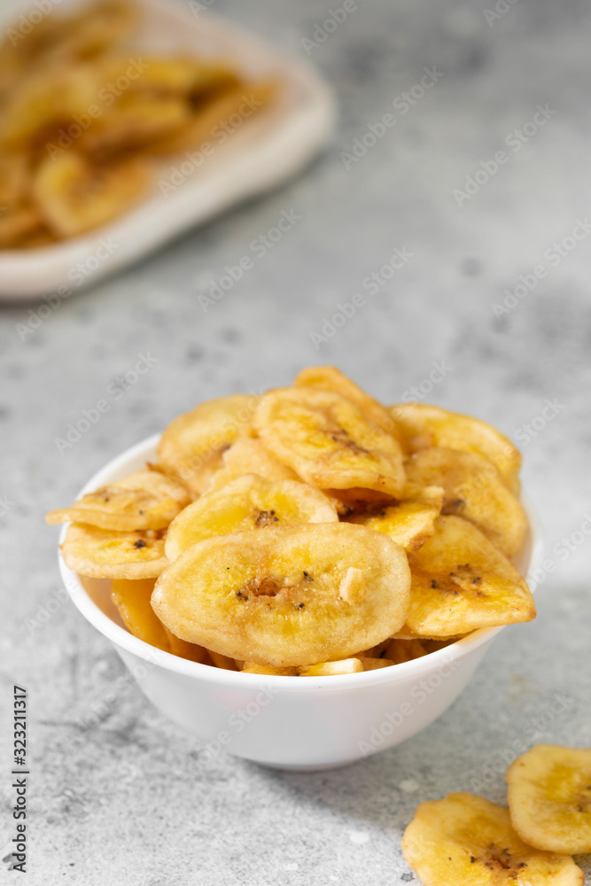 Dried bananas in a white ceramic bowl on a gray kitchen table. Banana chips. Vegetarian snack for proper nutrition Dried bananas in a white ceramic bowl on a gray kitchen table. Banana chips. Vegetarian snack for proper nutrition