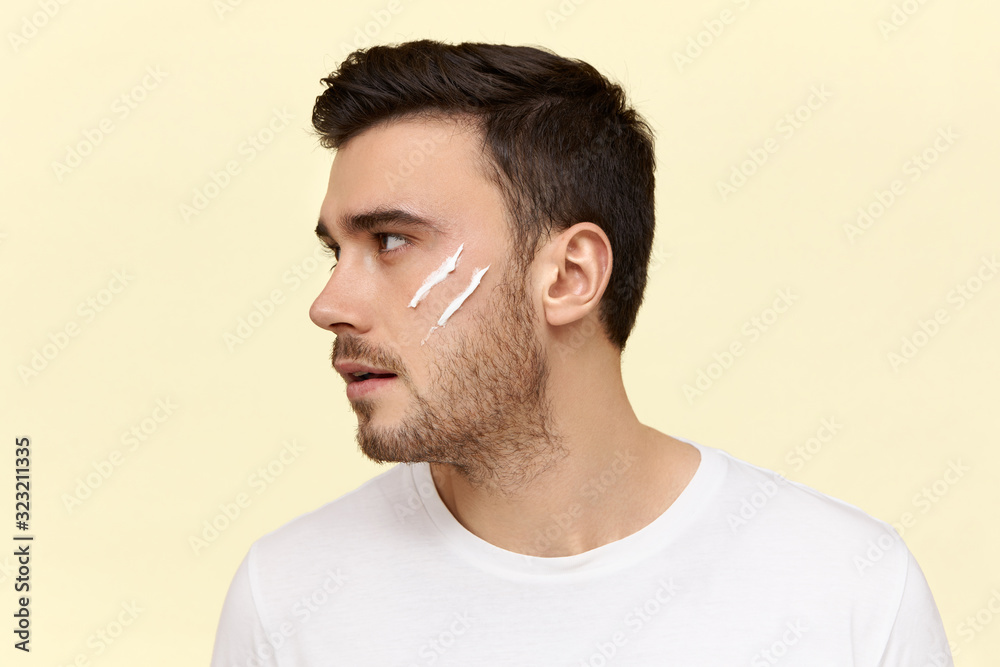 Obraz premium Profile shot of confident good looking young European guy with stylish hairdo and bristle applying moisturizer in front of mirror in the morning before work. People, beauty and care concept