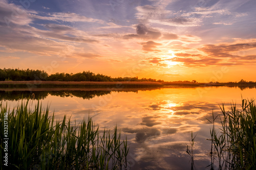 Fototapeta Scenic view of beautiful sunset or sunrise above the pond or lake at spring or early summer evening with cloudy sky background and reed grass at foreground. Landscape. Water reflection.