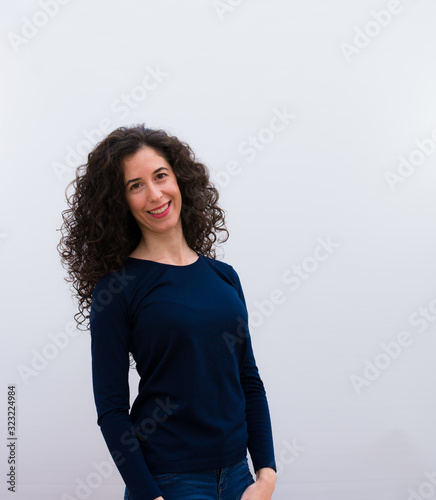young pretty woman with black curly hair looking happy, cheerful and confident, smiling proudly and looking to side against white wall.
