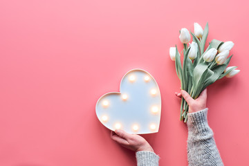 Springtime flat lay on pink, female woman hands holding bunch of white tulips, Spring flowers. Lightboard in heart shape in hands. Flat lay, top view, Mothers day or Valentine.