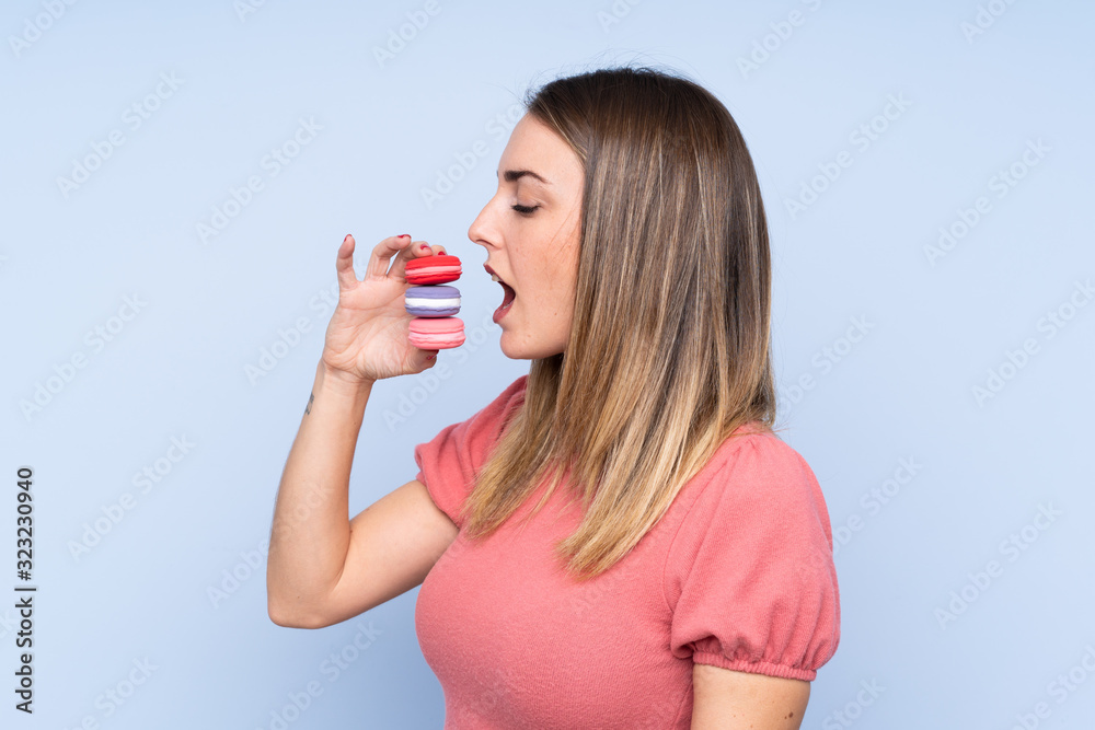 Young blonde woman over isolated blue background holding colorful French macarons and eating it Young blonde woman over isolated blue background holding colorful French macarons and eating it