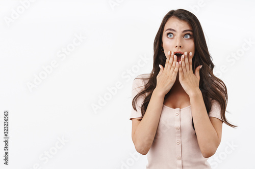 Astonished, startled attractive caucasian female in dress, gasping drop jaw, cover opened mouth from surprised, seeing something stunning and unbelievable, look upper left corner, white background