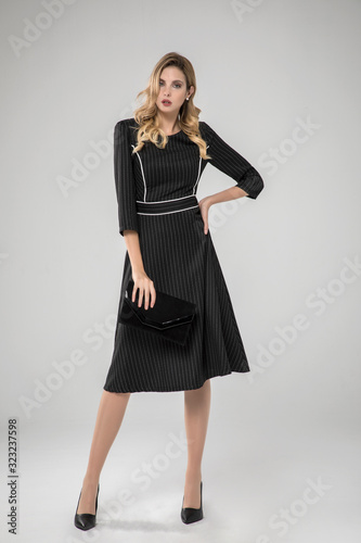 sexy charming model posing in white striped long black chic dress. black purse. white background. standing. looking at camera. studio shot.