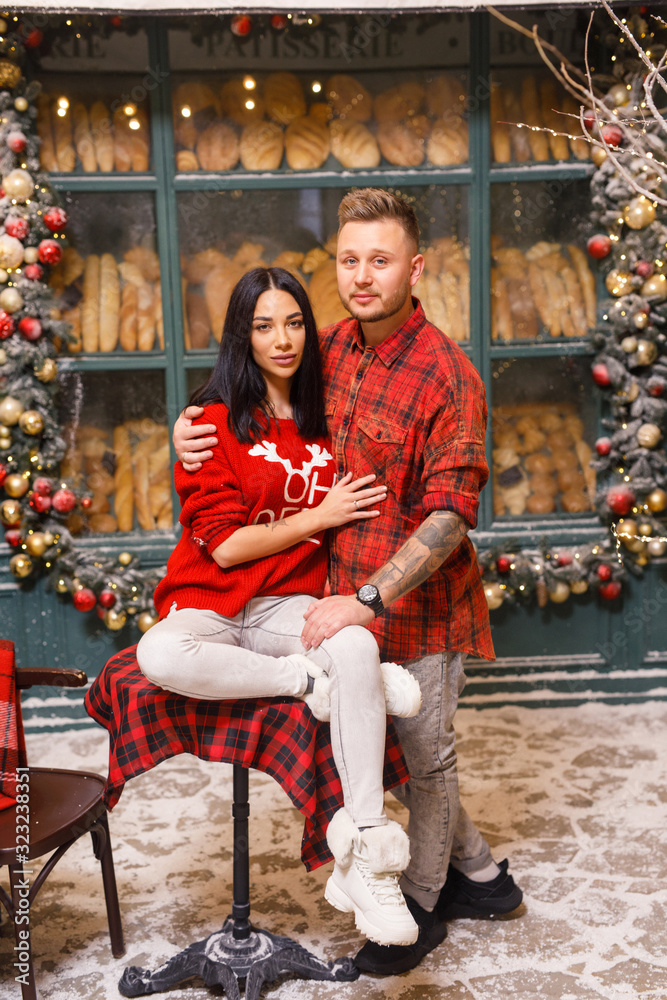 Young beautiful couple, a brunette girl in a red sweater and white pants, a man in a red shirt are sitting on the streets of Paris against the background of a window of a bakery shop Young beautiful couple, a brunette girl in a red sweater and white pants, a man in a red shirt are sitting on the streets of Paris against the background of a window of a bakery shop