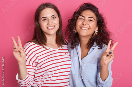 Indoor shot of two cheerful women friends posing isolated over pink background in studio, attractive brunette girls shoving v sign, posing with victory gesture, looking smiling directly at camera.