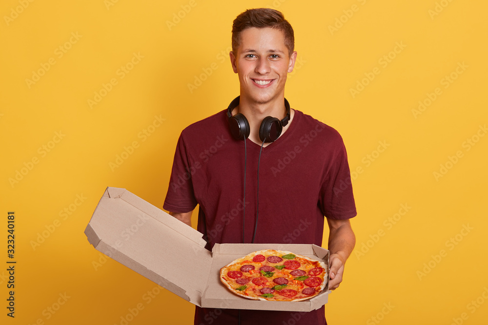 Indeoor of magnetic young boy holding opened box with pizza, having headphones around neck, wearing burgundy t shirt, male posing isolated over yellow background in studio. Delivery concept. Indeoor of magnetic young boy holding opened box with pizza, having headphones around neck, wearing burgundy t shirt, male posing isolated over yellow background in studio. Delivery concept.