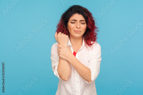 Hand injury. Office worker with fancy red hair in white shirt standing with grimace of pain, holding sore wrist, suffering carpal tunnel syndrome. indoor studio shot isolated on blue background © khosrork