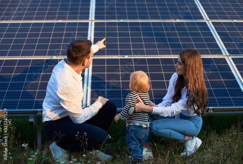 Back view of young family of three crouching near photovoltaic solar panel, getting acquainted with alternative energy, modern family concept