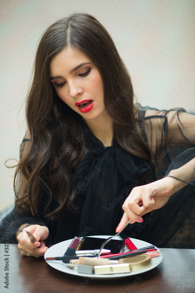 beautiful young woman in a black dress with makeup sits at a table with cosmetics in a plate beautiful young woman in a black dress with makeup sits at a table with cosmetics in a plate