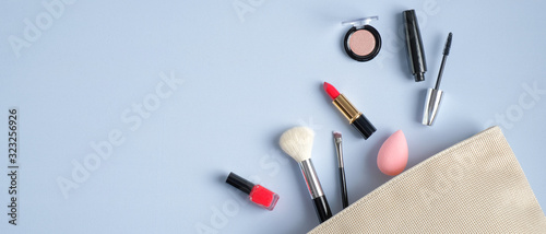 Cosmetic bag and makeup beauty products on blue background with copy space. Top view make-up brush, lipstick, mascara, eyeshadow spilling out of makeup artist pouch. Beauty salon banner design