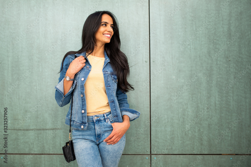 beautiful young arab woman with purse smiling by green wall looking away beautiful young arab woman with purse smiling by green wall looking away